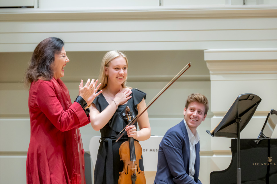 A female teacher and student, with one holding their violin, talking to each other and looking at a music sheet, on a well lit stage, with the male pianist, sitting next to the piano, listening to the